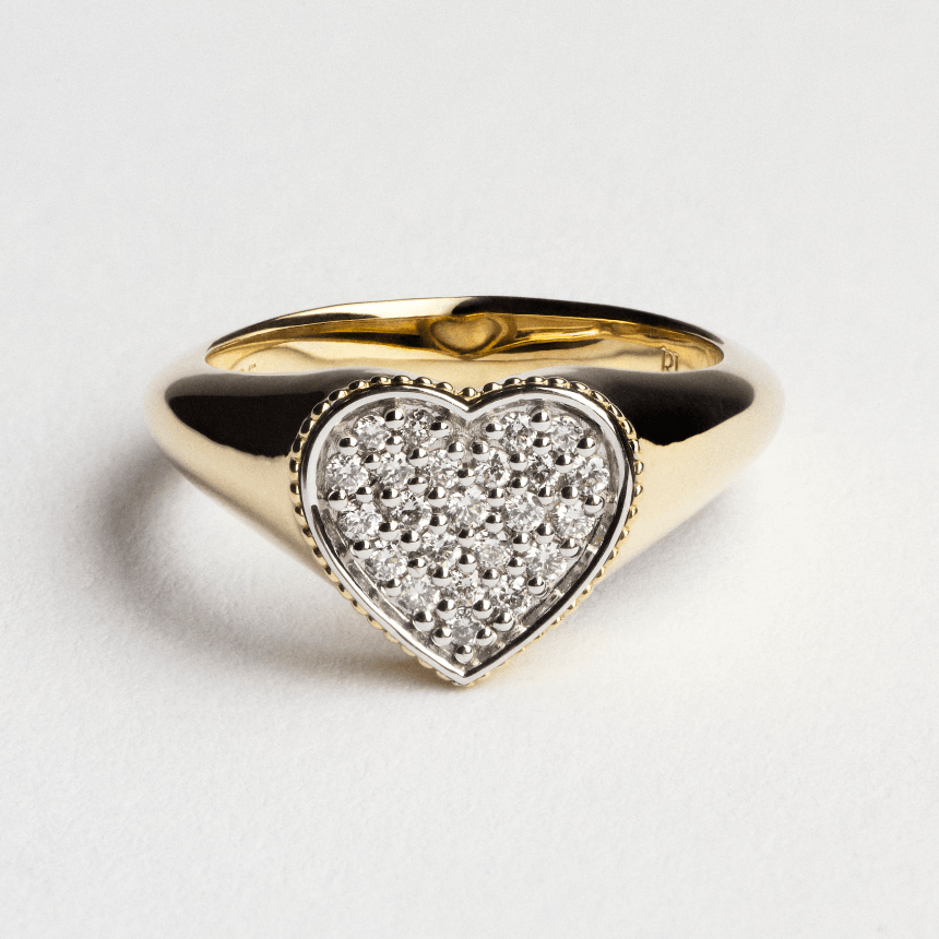 Gold pinky signet ring with a heart-shaped pave diamonds on a light gray background