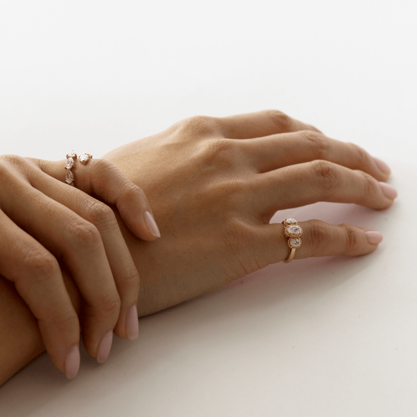 Two hands wearing gold diamond oval trilogy and pear cut rings on a light background